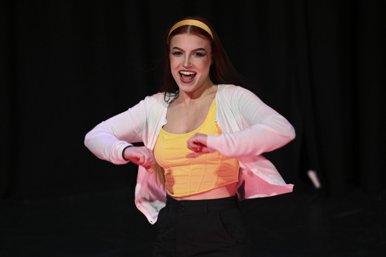 girl in yellow singlet, black pants and white cardigan dancing on stage.