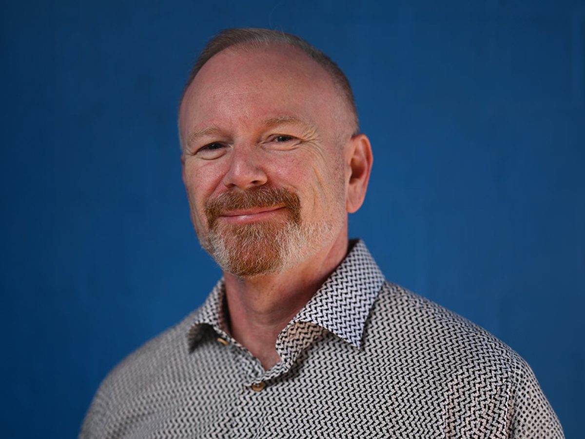 A man with a beard in a black and white shirt standing in front of a blue background