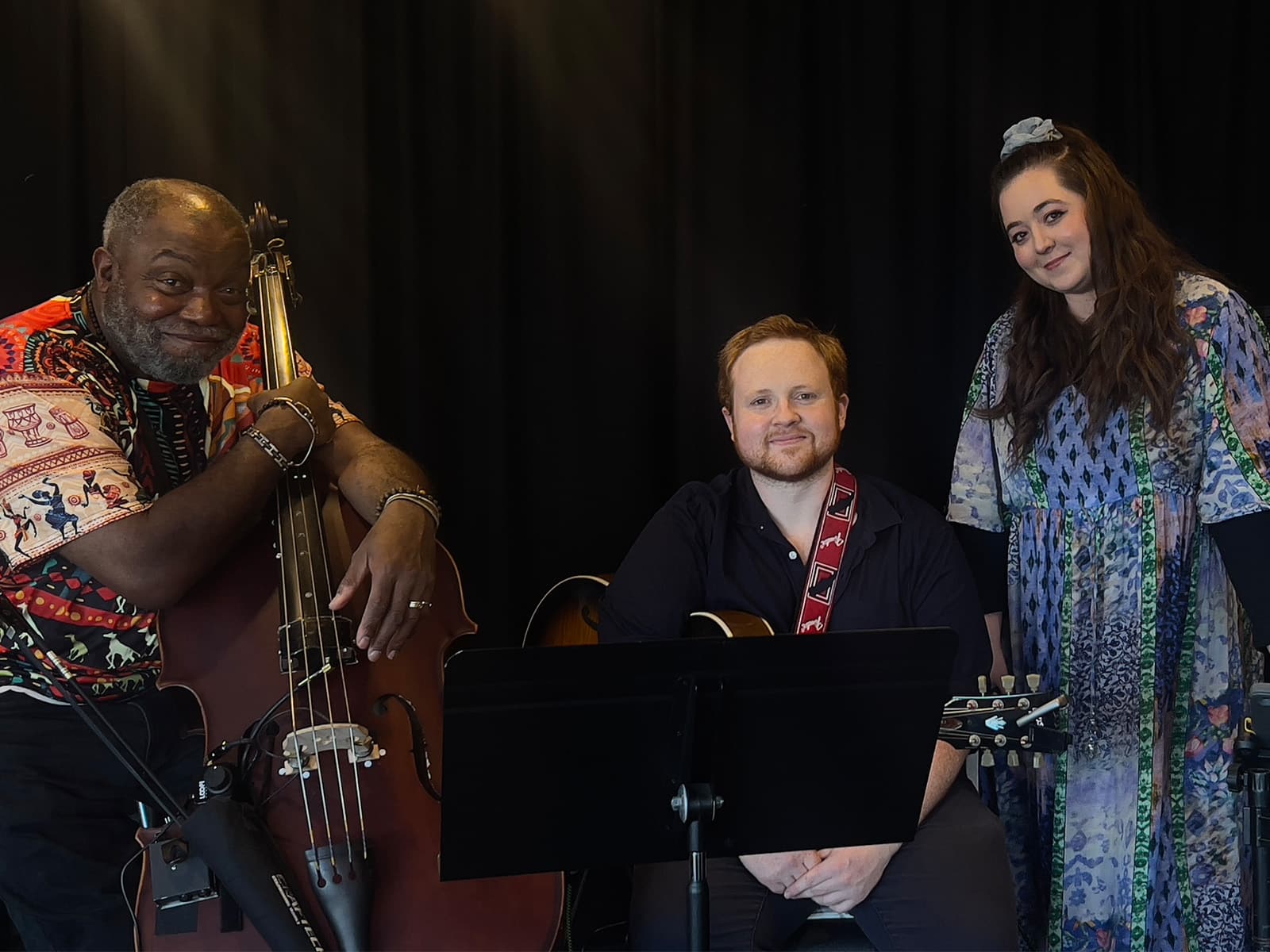 On the left is a black man holding a double bass, in the centre is a man with ginger hair and a beard holding a guitar and on the right is a woman with long dark hair in a colourful dress.