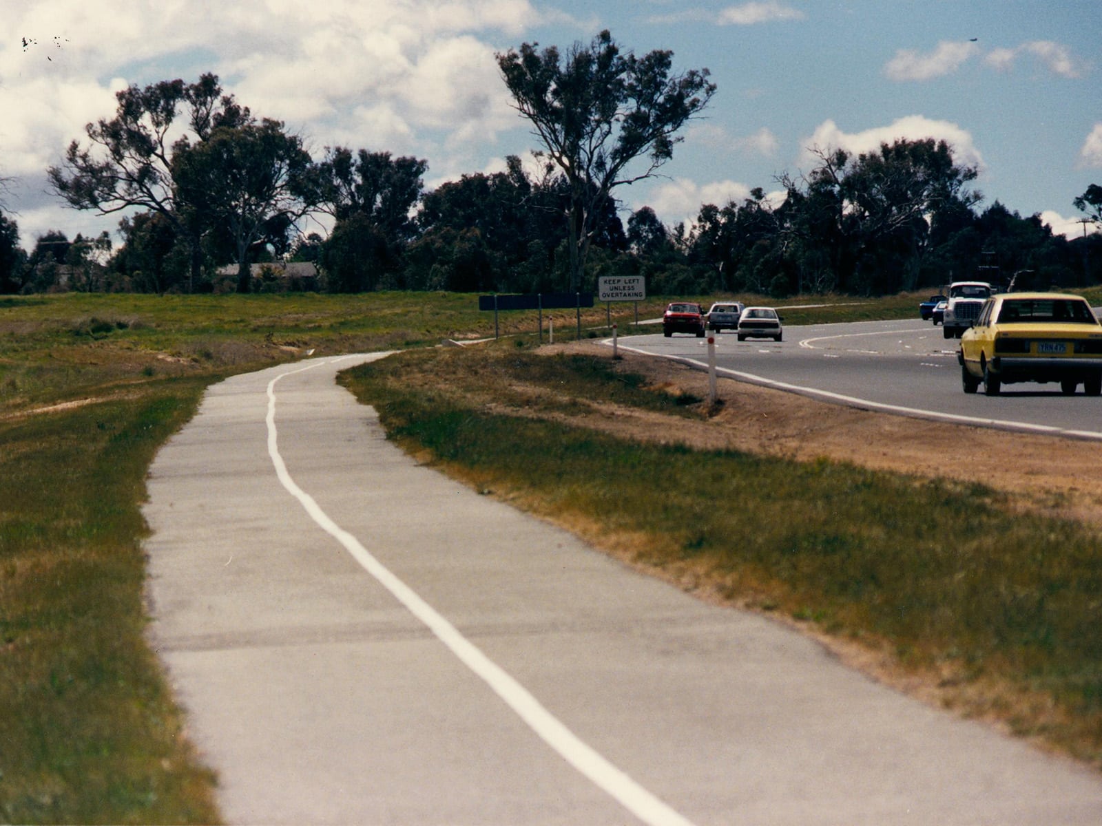 An image from the 1980s of a busy road, a bike path and grass under a blue sky