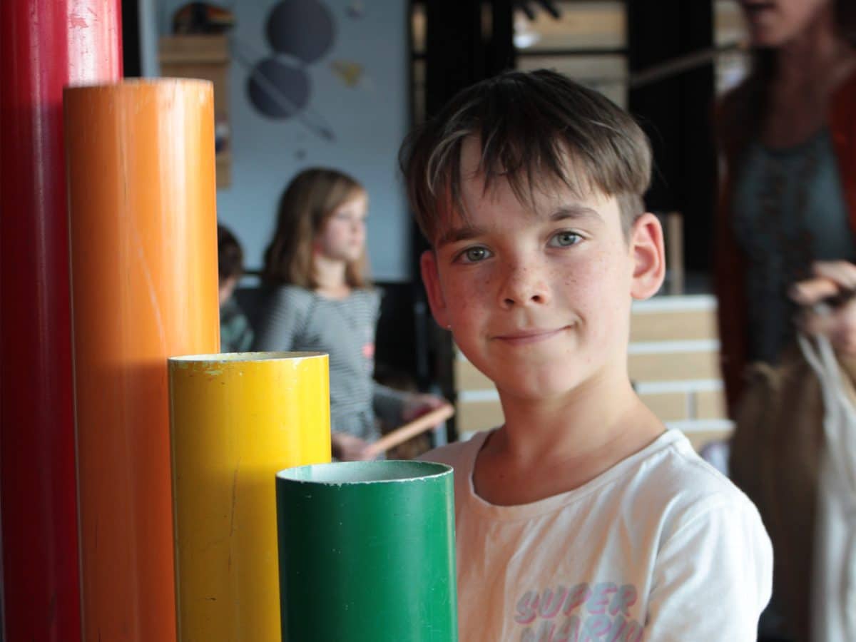 A young boy with dark hair and freckles stands next to rainbow coloured tubes.