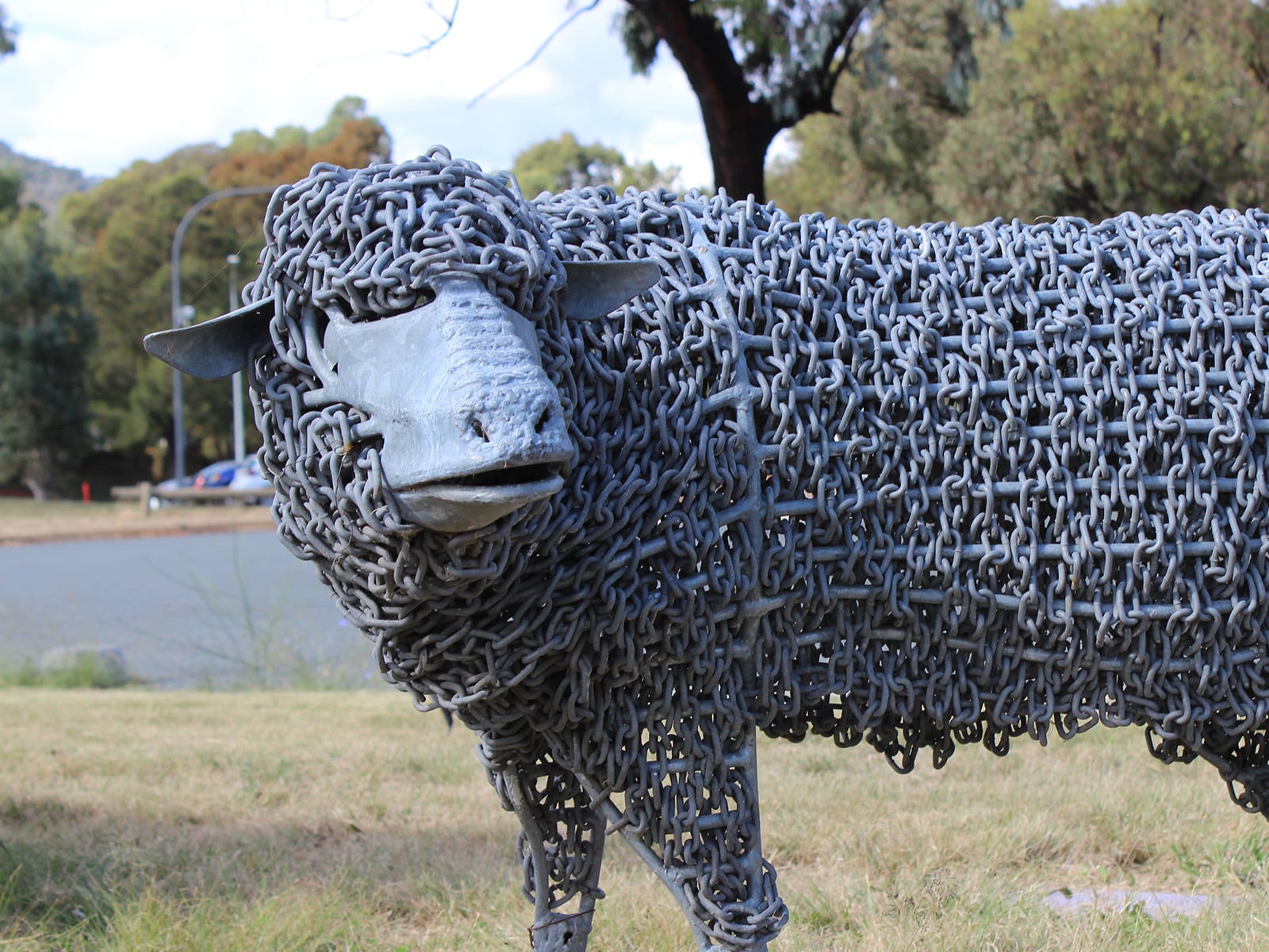 A photograph of a metal sheep sculpture standing on a green patch in front of a road.