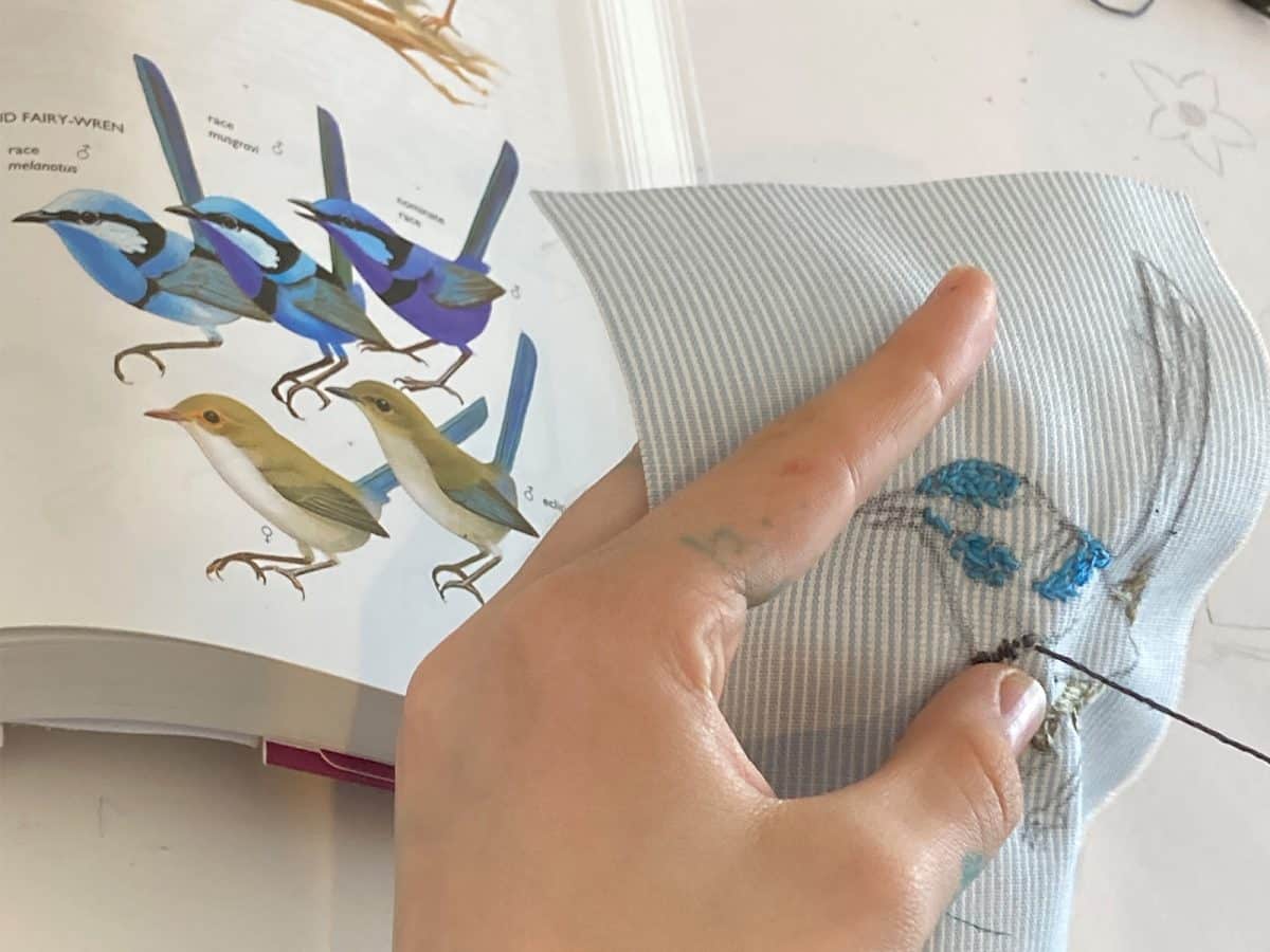 A hand holding blue and white striped fabric making with had stitching on it. A book open to illustrations of fairy wrens is in the background