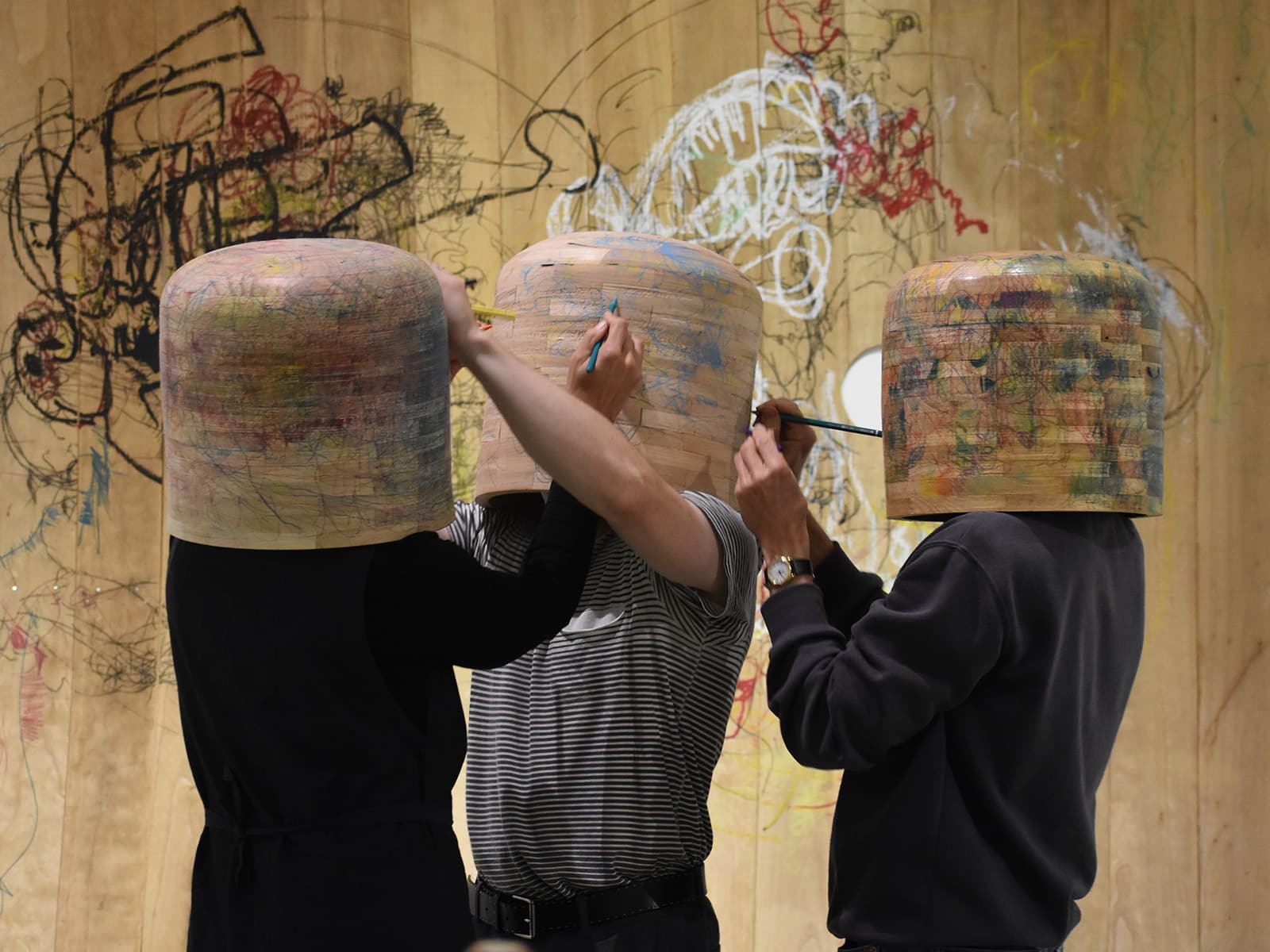 Three people with large wooden bowls on their heads draw on each other with coloured pencils