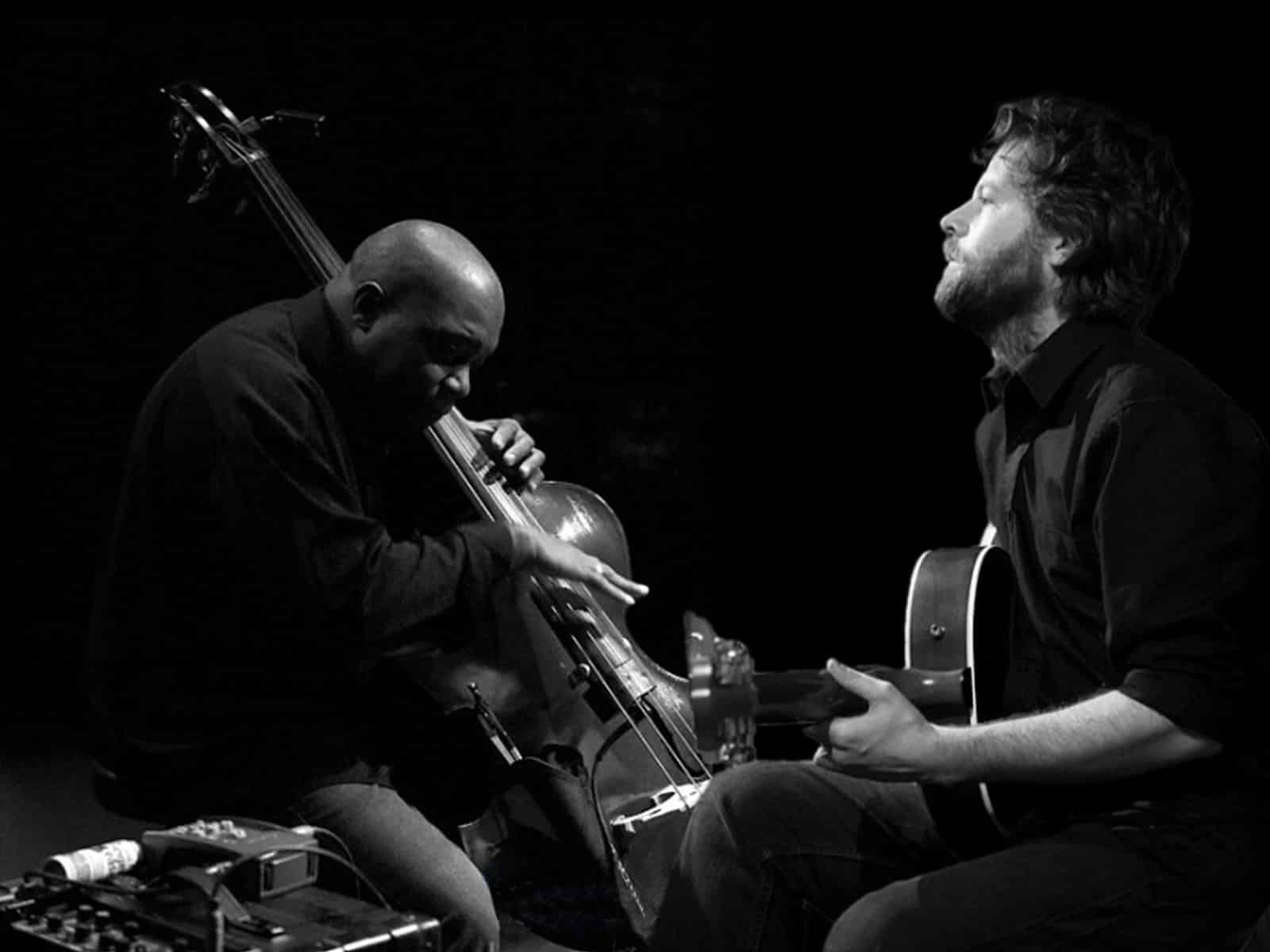 A black and white photo where a black man with a bald head plays the double bass and a white man with curly hair and a beard plays the acoustic guitar.