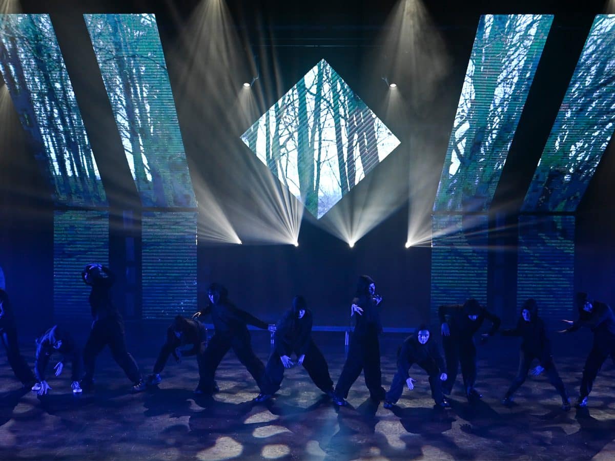Dancers dressed all in black perform in front of a projected image of a forest