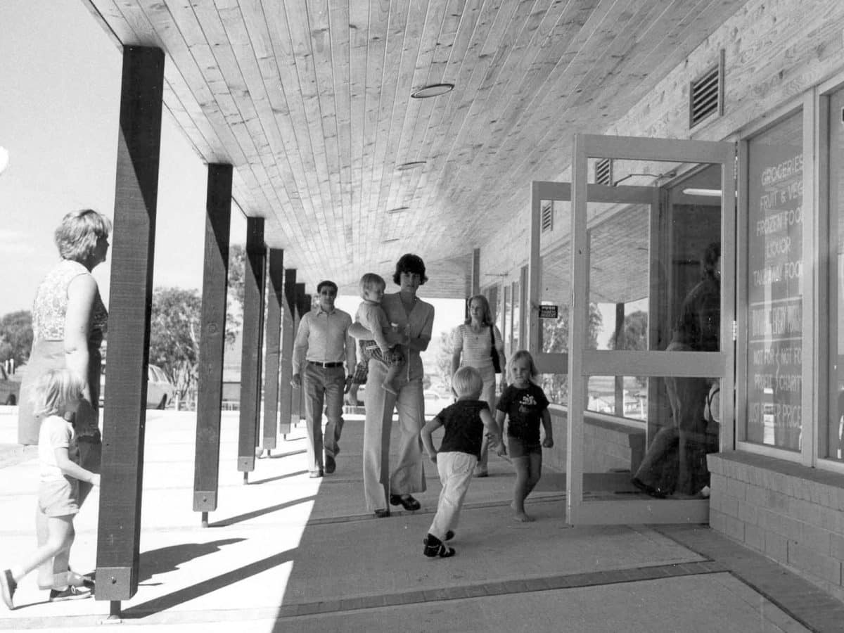 A black and white image from the 1970's of a group of adults and children walking into a local supermarket.