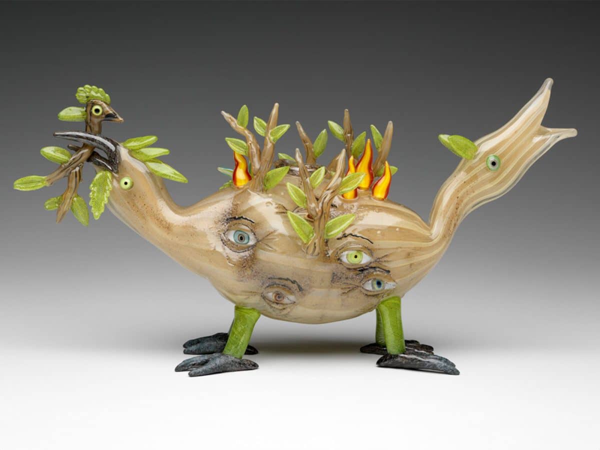 A glass creature with two heads, four legs, eyes in its body, branches and flames coming out of its body. One of the heads holds a stick creature in its beak.