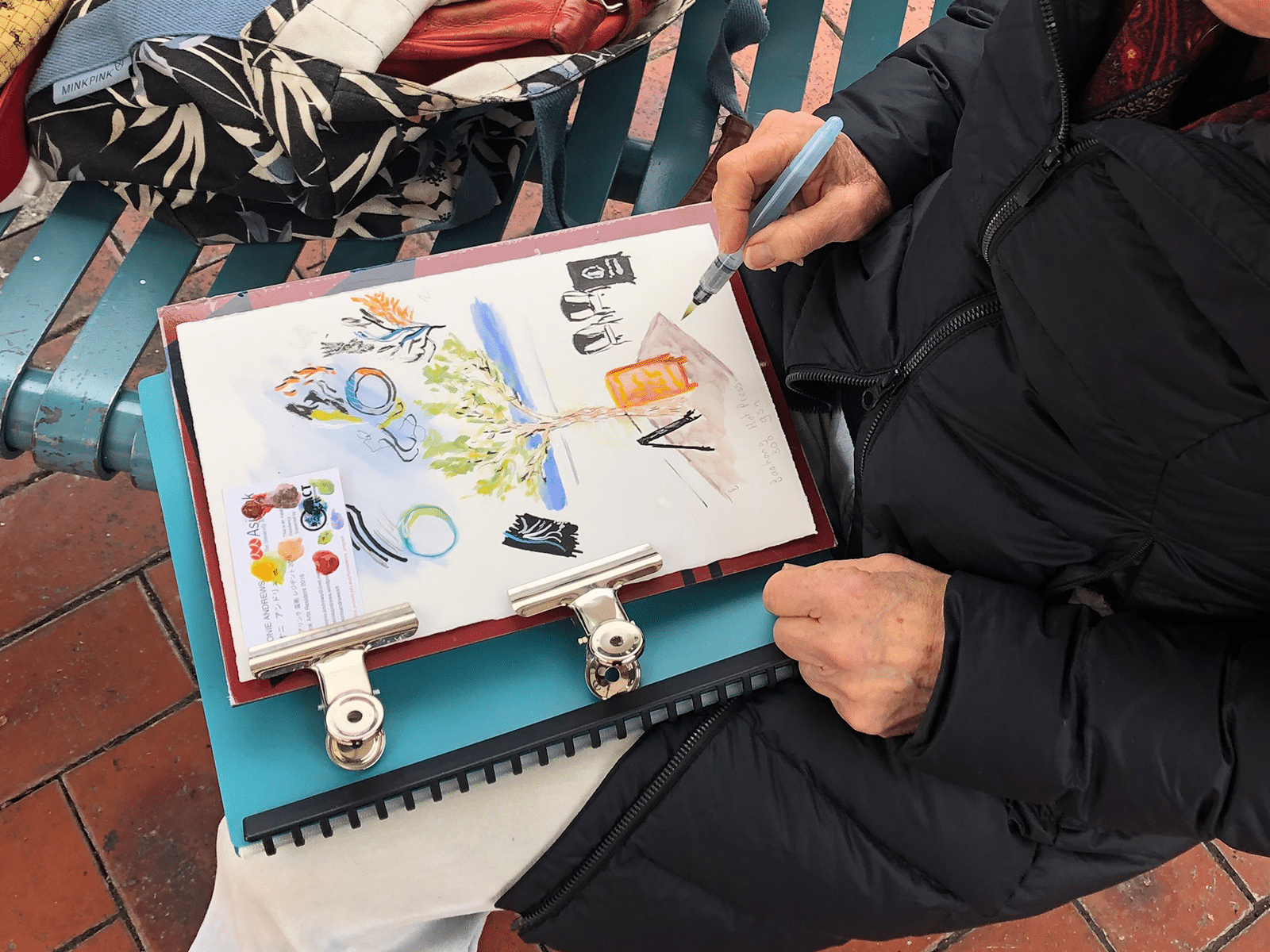 A person in a black puffy jacket paints a colourful image on a pad of paper on their lap.