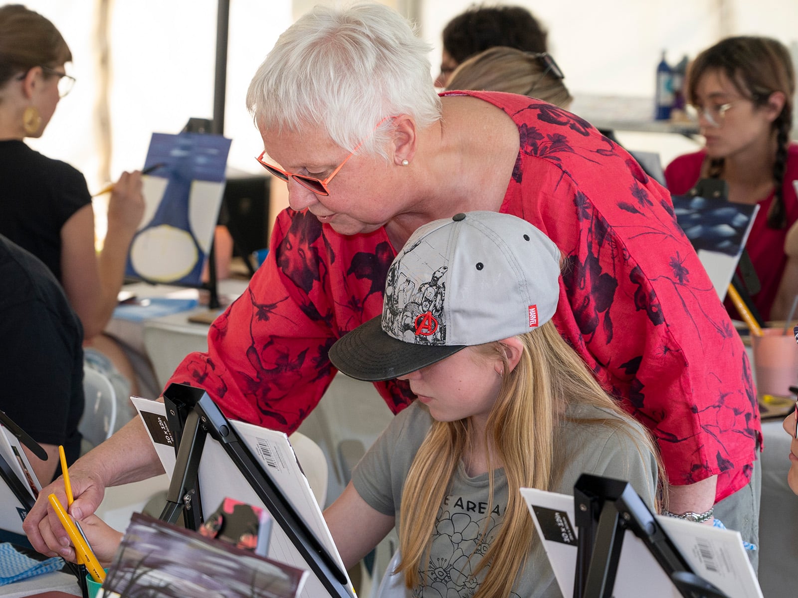 A woman with short white hair and orange glasses helps a young girl with long blond hair and a grey peaked cap do a painting