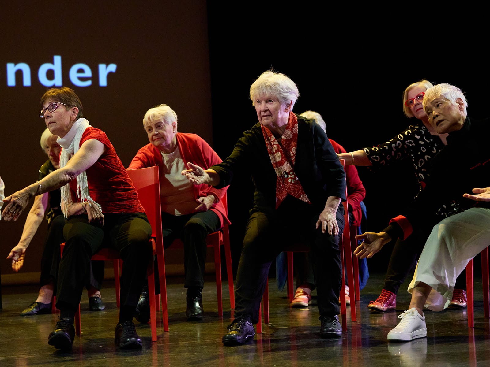 A group of older people are dancing while sitting in red chairs. They are all reaching towards to left of the image