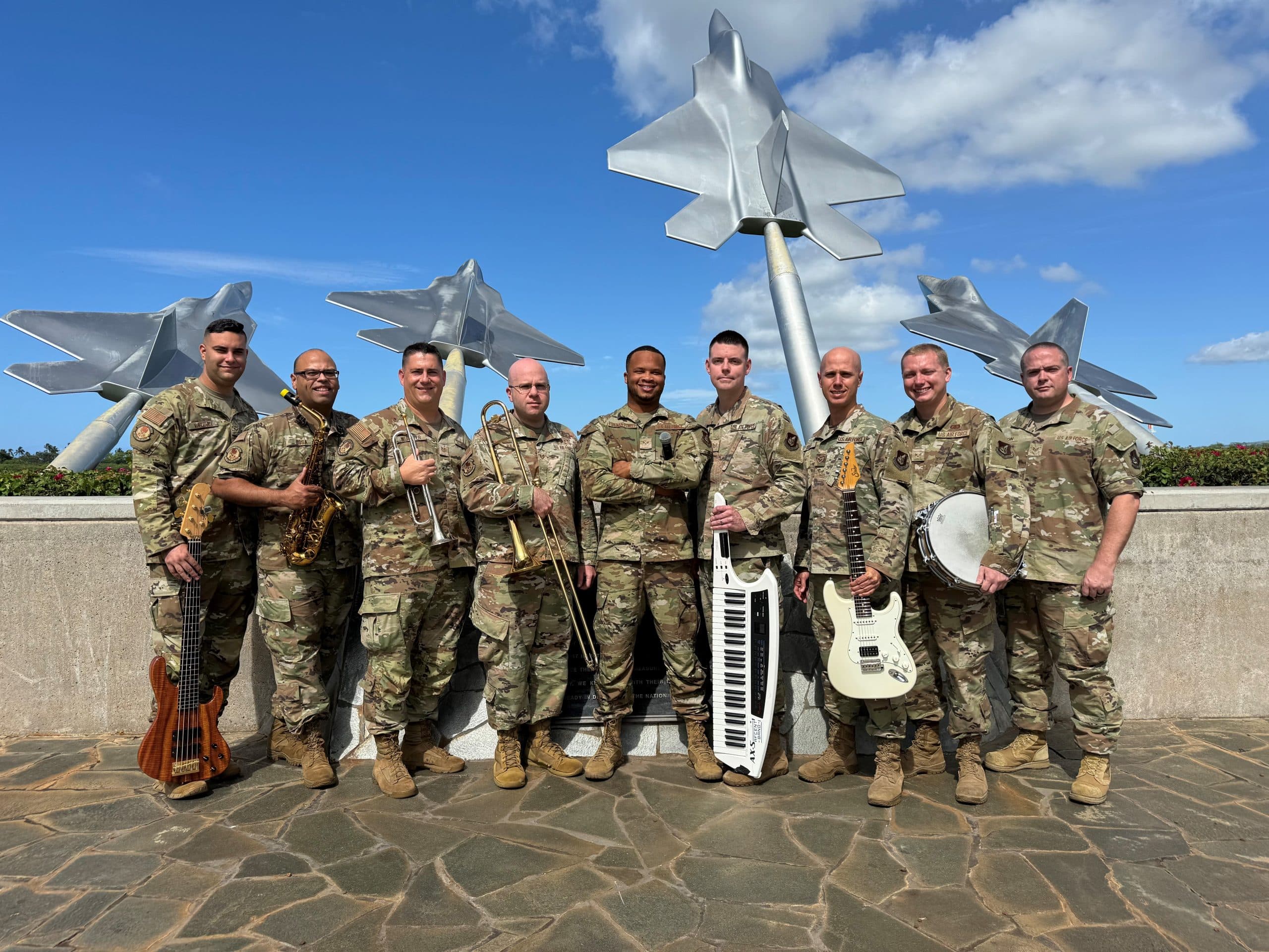 9 band members holding their instruments in camouflage, with sculptures of planes in the blue sky in the background